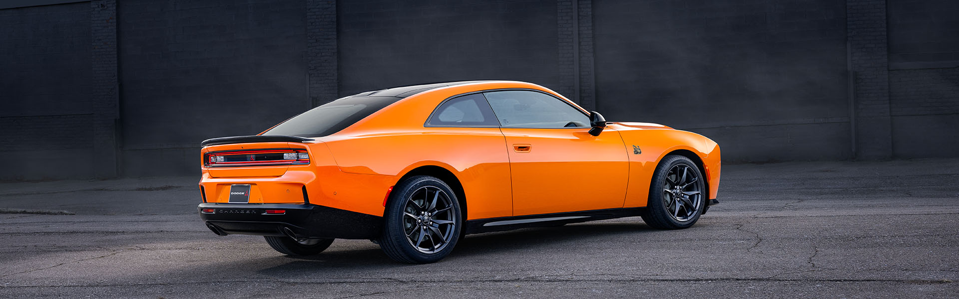 An orange Dodge Charger coupe parked on asphalt, showcasing its rear three-quarter profile against a dark, textured wall.